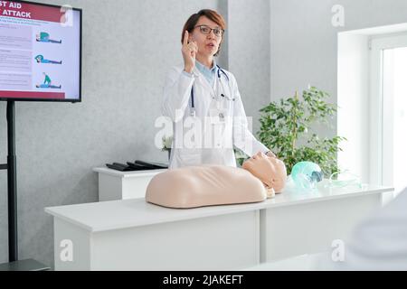 Reife Ärztin spricht über die Regeln der ersten Hilfe mit Schaufensterpuppe während des medizinischen Trainings Stockfoto