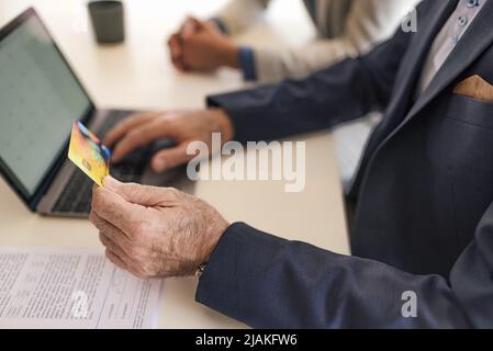 Leitender Geschäftsmann, der online über einen Laptop bezahlt. Ein männlicher Profi sitzt mit einer Kollegin zusammen. Der Schwerpunkt liegt auf der Kreditkarte im Büro. Stockfoto