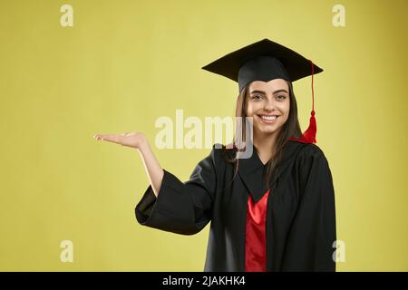 Vorderansicht eines hübschen Mädchens, das sein Studium an der Universität abschloss. Schöne weibliche stehend, lächelnd, Blick auf die Kamera, trägt Mortarboard und Graduate Kittel, Konzept der Jugend. Stockfoto