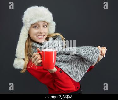 Attraktives Mädchen in einem Winterhut und einem grauen Schal hält eine rote Tasse in den Händen. Fotosession im Studio auf dunklem Hintergrund Stockfoto