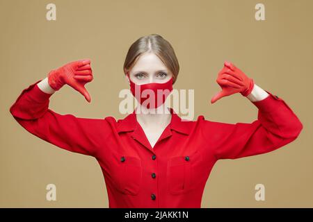 Blonde in roten Handschuhen und eine Maske zeigt ihren Daumen nach unten. Das Konzept der Prävention von Coronavirus covid 19. Stockfoto