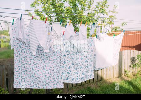 Saubere, gewaschene neugeborene Kleidung trocknet an sonnigen Tagen an einer Wäscheleine im Garten in der Nähe des Zauns. Stockfoto