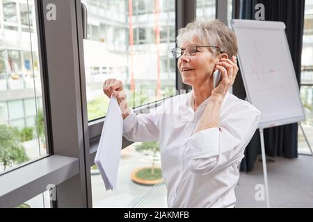Ältere Geschäftsfrau im Büro, die Dokumente hält und am Handy spricht Stockfoto