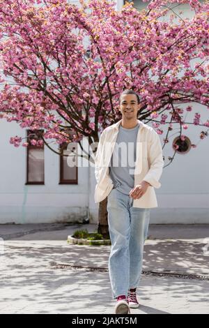 Lächelnder afroamerikanischer Mann in Hemdjacke, der mit der Hand in der Tasche in der Nähe des japanischen Kirschbaums läuft Stockfoto