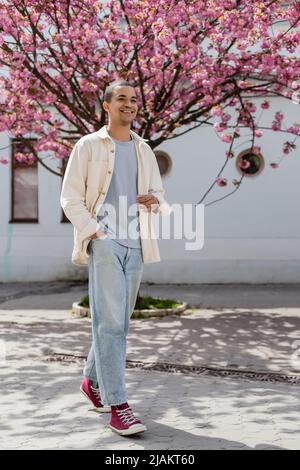 Glücklicher junger afroamerikanischer Mann in Hemdjacke, der mit der Hand in der Tasche in der Nähe von Kirschbaum läuft Stockfoto
