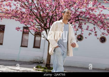 Positiver afroamerikanischer Mann in Hemdjacke, der mit der Hand in der Tasche in der Nähe von Kirschbaum läuft Stockfoto