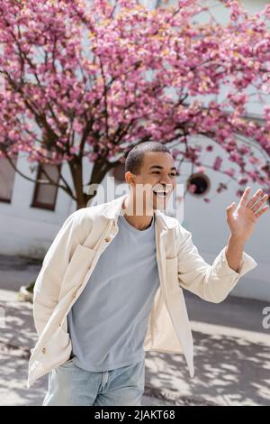 Junger afroamerikanischer Mann in Hemdjacke winkt Hand neben Kirschbaum Stockfoto