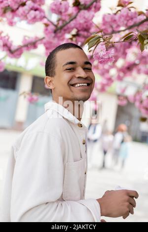 Positiver afroamerikanischer Mann, der in der Nähe des blühenden Kirschbaums lächelt Stockfoto