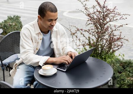 afroamerikanischer Mann mit Laptop in der Nähe einer Tasse Latte mit Schaumstoff auf dem Tisch auf der Café-Terrasse Stockfoto