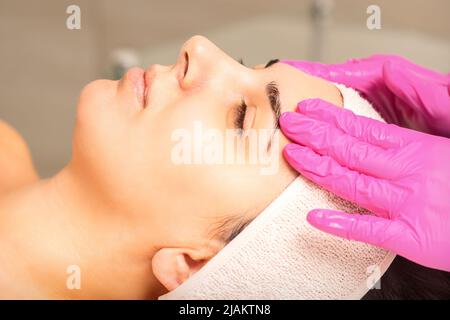 Der Kosmetikerin mit Handschuhen trägt eine Feuchtigkeitsmaske mit Peeling-Creme auf das weibliche Gesicht auf. Gesichtskosmetologie Behandlung. Verfahren für die Gesichtspflege Stockfoto