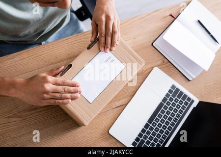 Beschnittene Ansicht des afroamerikanischen Verkäufers, der im Geschäft einen Karton mit Versandetikett in der Nähe von Laptop und Notebook hält Stockfoto