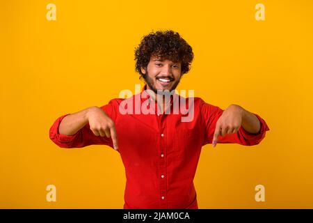 Hier und jetzt. Bärtiger, hübscher Mann, der mit dem Finger nach unten zeigt und mit glücklichem Gesichtsausdruck auf die Kamera blickt. Innenaufnahme des Studios isoliert auf orangefarbenem Hintergrund Stockfoto