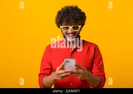 Porträt von fröhlichen Brünette Mann mit Bart mit Handy und lächeln, lesen gute Nachricht, genießen mobile Anwendung. Innenaufnahme des Studios isoliert auf orangefarbenem Hintergrund Stockfoto