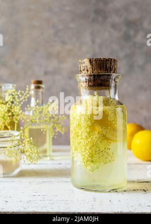 Holunderbeerenwasser oder Sirup in Glasflasche mit frischen Blumen und Zitronen. Grauer und weißer Hintergrund Stockfoto