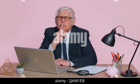 Shh be Ruhe bitte. Leitender Geschäftsmann in Anzug drückt Zeigefinger auf die Lippen macht Stille Geste Zeichen nicht erzählt Geheimnis sitzt am Arbeitsplatz Schreibtisch im Büro. Älterer Mann auf rosa Studio Wand Hintergrund Stockfoto