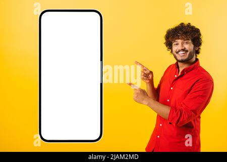 Spielerisch schöner lockiger indischer bärtiger Mann zeigt die Finger auf riesiges Smartphone mit leerem leeren Bildschirm isoliert auf gelbem, glücklichem lateinischem Kerl präsentiert mobile App, zeigt Best Deal, Copy Space, Vorlage Stockfoto