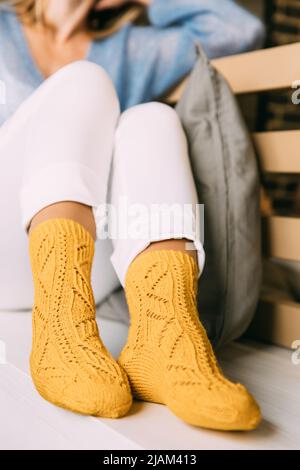 Nahaufnahme einer gemütlichen Frau in warmen gelben gestrickten Wintersocken, weißen Hosen und blauem warmen Pullover, die im Winter auf einer Bank im Café sitzen Stockfoto