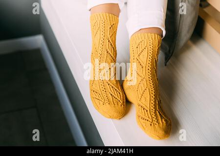 Nahaufnahme von weiblichen Beinen in gelben gestrickten Wintersocken, weiße Hosen, die im Winter auf einer Bank im Café sitzen. Fauler Tag in gestrickten Socken an Stockfoto