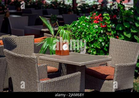 Rattan-Kunststoffarmaturen auf der sonnenbeleuchteten Terrasse des Straßenrestaurants, quadratischer Tisch mit Serviettenhalter und Stühlen mit weichen Kissen in der Nähe grüner p Stockfoto