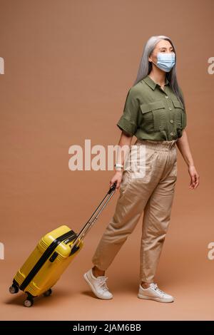 Reife Frau in Gesichtsmaske ist mit Koffer isoliert zu Fuß. Isolierte Dame mit einem Koffer geht mit ihr in Gesichtsmaske und schaut in die Kamera. Frau in Gesichtsmaske geht mit Flugtasche Stockfoto
