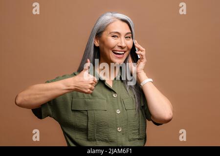 Fröhlich lächelnde ältere grauhaarige Geschäftsfrau, die auf dem Smartphone spricht und der Kamera auf braunem Hintergrund isoliert den großen Finger zeigt. Reife Dame hat angenehme Telefongespräche Stockfoto