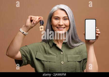Portrait of adorable Asian Woman Holding a car key, presenting smart phone isoliert auf braunem Hintergrund in Studio mit Copy Space Stockfoto