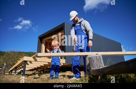 Vater mit Kleinkind Sohn Gebäude Holzrahmen Haus. Männliche Bauherren, die auf der Baustelle Nagel in die Planke schlagen, tragen an sonnigen Tagen Helm und blaue Overalls. Zimmerei- und Familienkonzept. Stockfoto