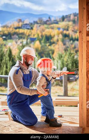 Vater mit Kleinkind Sohn Gebäude Holzrahmen Haus. Junge hilft seinem Vater, schlägt Nagel auf die Planke auf der Baustelle, trägt Helm und blaue Overalls an sonnigen Tagen. Zimmerei- und Familienkonzept. Stockfoto