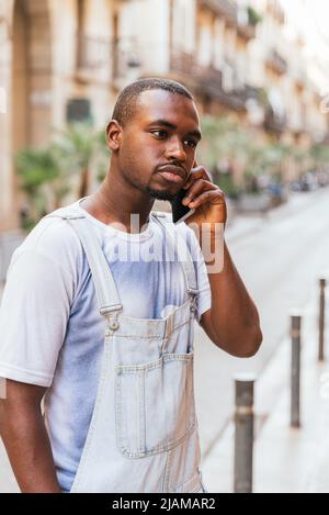 Stylischer junger ernsthafter afroamerikanischer Kerl im trendigen Outfit mit dunklen Haaren und Barthaar. Er spricht auf dem Handy, während er an einem sonnigen Tag auf der Stadtstraße steht Stockfoto