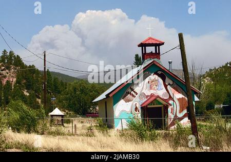 In den Bergen in der Nähe von Las Vegas, NM, steht eine Kirche, die im 17. Jahrhundert von den Vorfahren der Bewohner von Montezuma, New Mexico, erbaut wurde. 5.Mai 2022. Hinter der Kirche, El Cristo Ray, wölben sich Wolken vom Waldfeuer des Calf Canyon/Hermits Peak in den Himmel. Das Feuer hat bereits zahlreiche Kulturstätten zerstört, die für die hispanische Bevölkerung von Bedeutung sind. Die Bewohner pflegen die Kirche, während sie nur für besondere Anlässe genutzt wird. Die kombinierten Brände kamen in die Nähe der benachbarten Stadt Las Vegas im Norden von New Mexico, bevor sie von Feuerwehrleuten gestoppt wurden. Das Waldfeuer auf dem Hermits Peak war zu sehen Stockfoto