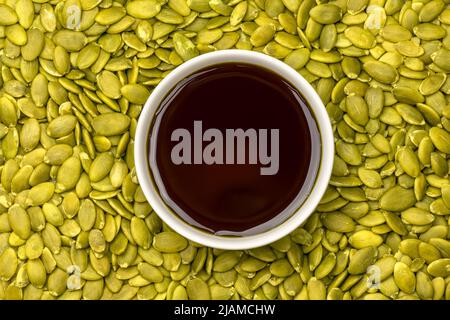 Kürbisöl in weißer Schale auf Hintergrund geschälter roher Kürbiskerne Draufsicht. Stockfoto