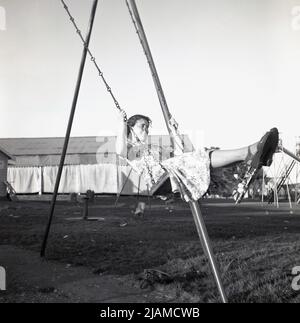 1950s, historisch, draußen auf einem Spielplatz am Meer, eine Dame in einem blumigen Kleid der Zeit, auf einer Schaukel, die an den Metallketten hält, zeigt gute Technik, Beine tgethering, Füße nach oben zeigend, England, VEREINIGTES KÖNIGREICH. Stockfoto