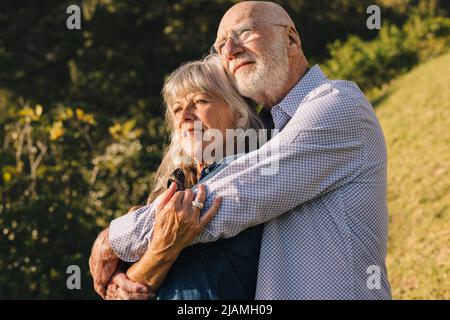 Romantisches Seniorenpaar umarmt sich gegenseitig, während es in einem Park steht. Ältere Paare, die sich gegenseitig die Sonne in den Armen genießen. Reifes Paar, das Qua ausgibt Stockfoto