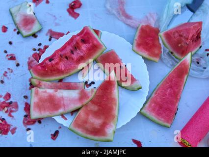 Teile der Wassermelone, die von Kindern gegessen wurden, wurden auf einem Teller und einem weißen Tisch vermasselt. Alles schmutzig mit Saft und Wassermelonen Samen im Freien. Portionen Wasser Stockfoto