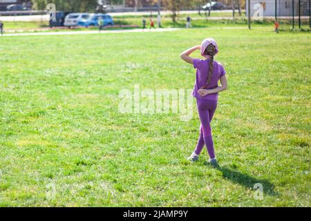 Ein Kind auf einem grünen Rasen blickt in die Ferne. Das Mädchen sucht nach ihren Eltern. Ein Schulmädchen schaut zu. Mädchen im Sommer im Park. Stockfoto