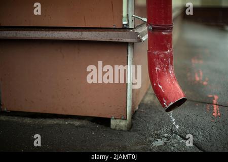 Downspout im Gebäude. Wasser fließt vom Dach auf Asphalt. Rotes Rohr zum Ablassen von Sedimenten. Stockfoto