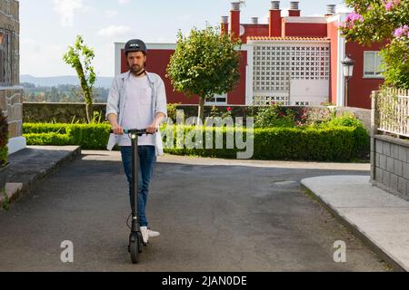 Bild eines jungen Mannes in legerer Kleidung, der auf einem Elektroroller auf der Straße steht Stockfoto