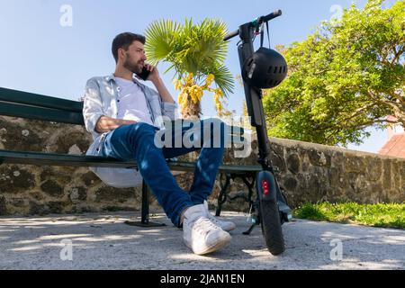 Der junge Mann spricht auf dem Smartphone auf der Bank neben seinem Elektroroller Stockfoto