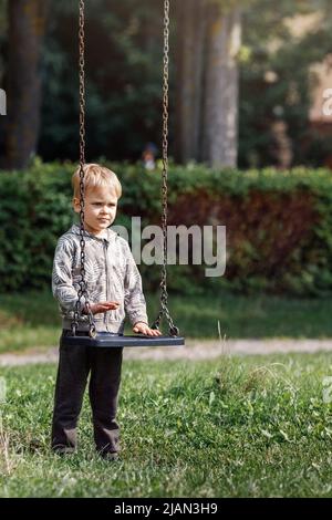 Ein attraktiver kleiner Junge steht neben einer Schaukel mit Ketten in einem Stadtpark, ein Kind macht sich bereit zum Schaukeln. Vertikales Foto. Stockfoto