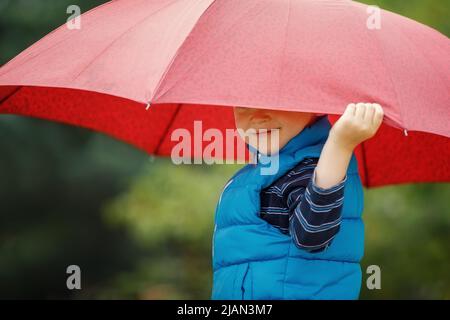 Glückliches Kind mit einem Regenschirm, der im Sommer draußen im Regen spielt. Der Junge lächelt und versteckt sein Gesicht unter einem Regenschirm. Stockfoto