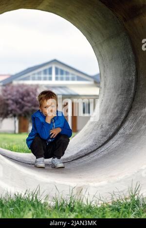 Der kleine Junge spielt im Hof, er driftet in ein großes Betonrohr und hockt zur Ruhe. Vertikales Foto. Stockfoto