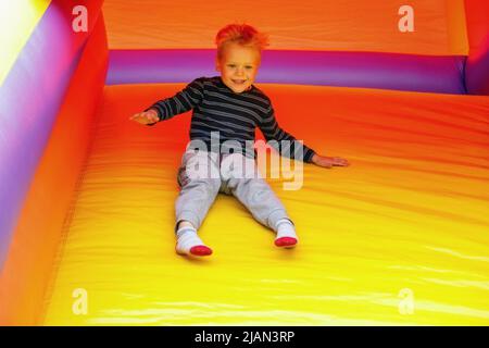 Ein fröhlicher, lachender Junge sitzt und rutscht auf einem leuchtend gelben Gummitrampolin hinunter. Bild in Bewegung, ein mutiges Kind hat vor nichts Angst. Stockfoto