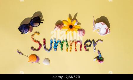 Die flache Laie der hellen Inschrift des Wortes Sommer aus den Süßigkeiten in Form von den Meereskieseln, den schönen Muscheln, der Blume, der Sonnenbrille auf dem gelben Hintergrund Stockfoto