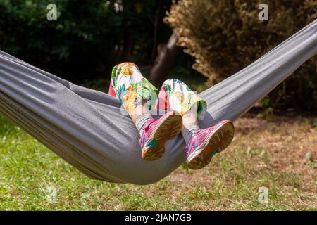 Anonymes Kind, Mädchen im Schulalter, das auf einer Hängematte liegt, die Füße nach oben, das Gesicht verdeckt, Szene im Freien, Sommerspaß und Freizeit, Ausruhen, Langeweile abstrakt Co Stockfoto