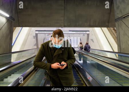 Junger Mann mit Gesichtsmaske aufgrund einer Coronavirus-Epidemie, der sein Telefon auf der Rolltreppe in der U-Bahn-Station ansah Stockfoto