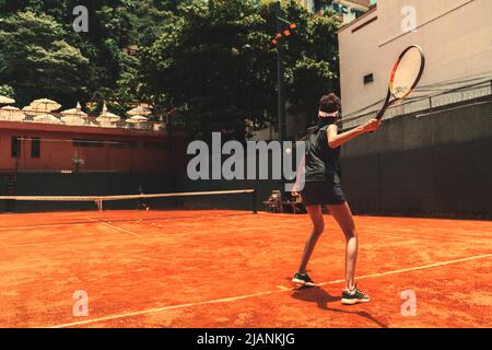 Blick von hinten auf eine schlanke, schnelle Sportlerin auf dem Tonfeld eines privaten Sportvereins während ihres Tennistrainings: Sie schwingt h Stockfoto