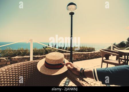 Blick auf die Hand eines eleganten schwarzen Mannes, der einen modisch zugeschnittenen marineblauen Anzug trägt und einen klassischen goldenen Strohhut hält, während er im Außenansicht sitzt Stockfoto