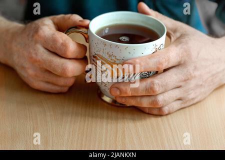 Männliche Hände mit Vitiligo-Verfärbung (fleckiger Verlust der Hautfarbe), halten eine vergoldete Tasse Hibiskus-Tee. Die Teeblätter stehen auf dem Tisch. Stockfoto