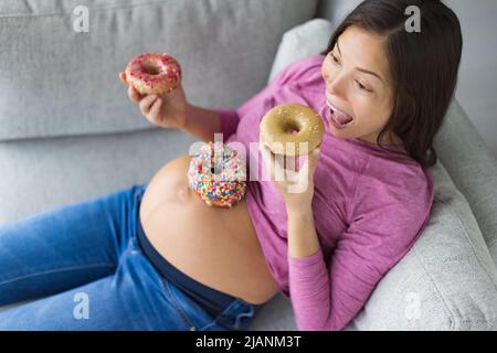Schwangerschaft Heißhunger lustige asiatische Schwangere Frau essen Donuts zu Hause hungrig nach Dessert-Kuchen und süßen Snacks. Gestationsdiabetes, Ernährung Stockfoto