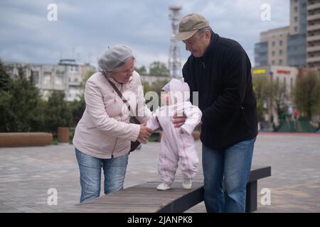 Kleines Mädchen, das mit Mann und Frau im Freien läuft. Großvater und Großmutter helfen der Enkelin beim Gehen Stockfoto